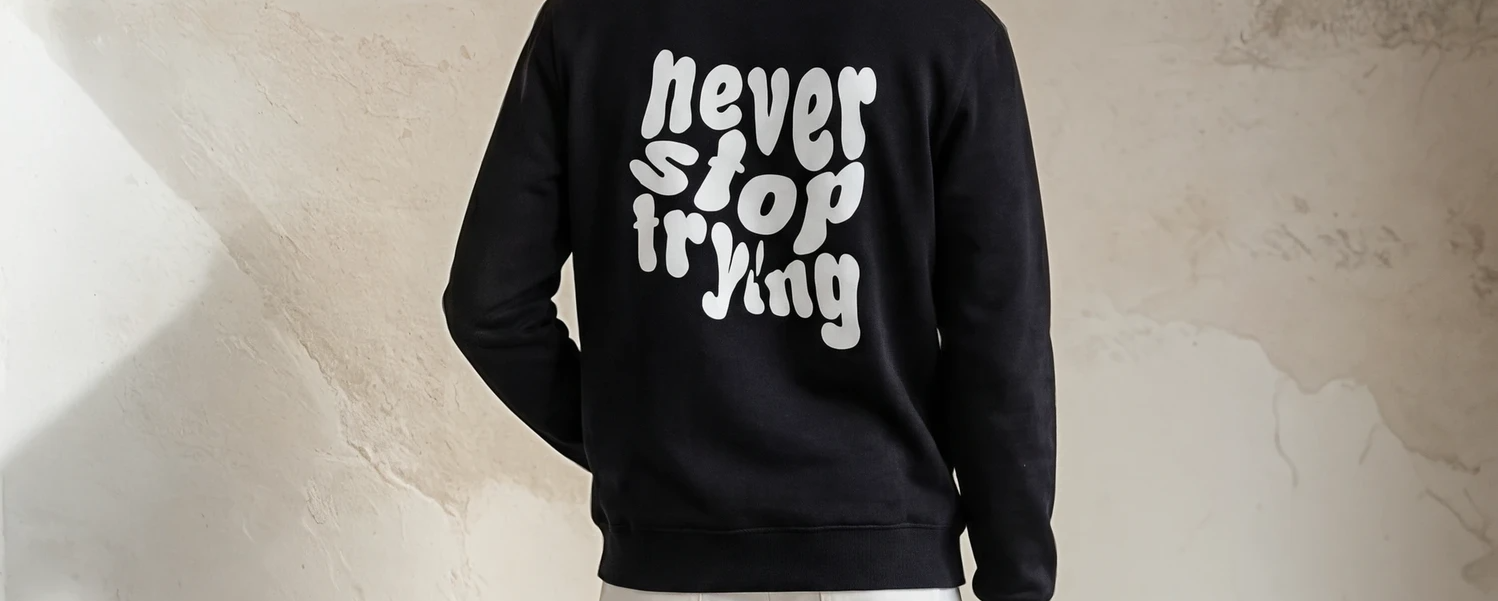 Black sweatshirt with 'never stop trying' text on the back against a beige wall.